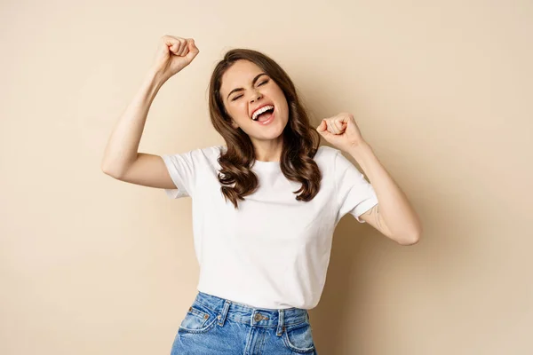 Enthusiastic adn surprised woman winning, dancing and celebrating, standing in tshirt over beige background — Foto de Stock