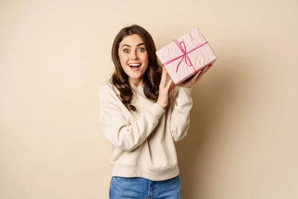 Emocionado linda chica sacudiendo caja con regalo, adivinando lo que está dentro presente, de pie feliz sobre fondo beige — Foto de Stock