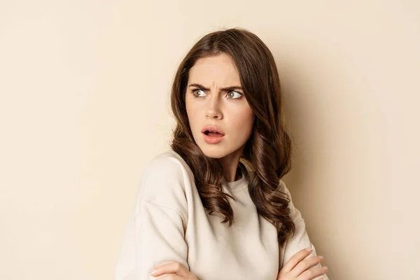 Close up of confused young woman, looking behind with skeptical shocked face expression, standing against beige background — Stock fotografie