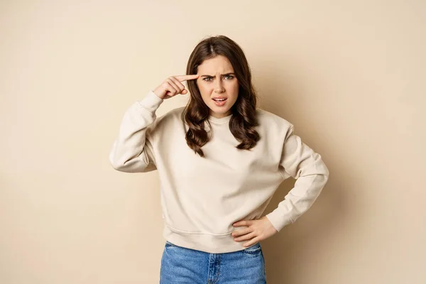 Frustrated woman roll finger near head and complaining, insulting someone, standing over beige background — Foto Stock