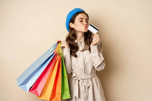 Portrait of cute happy girl kissing her credit card after buying gifts, holding shopping bags, standing over beige background — Stockfoto