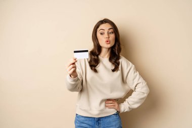 Smiling beautiful woman showing credit card, concept of shopping, contactless payment, standing over beige background