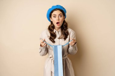 Image of girl looking surprised after opening gift bag, looking at present amazed, standing in stylish outfit over beige background