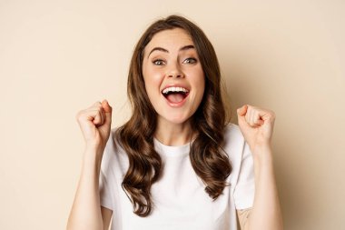 Close up of cheerful, happy beautiful woman winning, chanting and rejoicing, shouting supportive and looking hopeful at camera, yearning to win, beige background