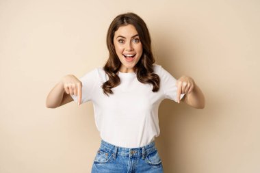 Smiling happy girl pointing fingers down and showing banner, company logo below, click link, standing over beige background