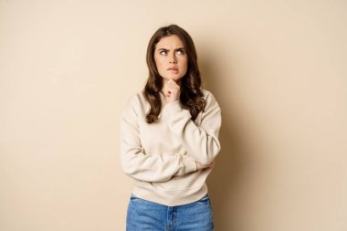 Image of beautiful woman with serious face thinking, standing over beige background