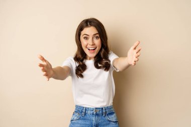 Gülümseyen mutlu genç bir kadının portresi, kucaklaşmak için kollarını uzatan, kucaklaşmak için ellerini uzatan, smth almak için uzanan, bej arka planda duran