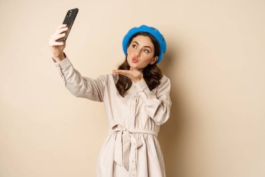 Beautiful modern woman posing for selfie, holding smartphone, taking picture photo on mobile phone, standing happy against beige background