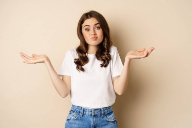 Young confused woman shrugging shoulders and with clueless face expression, standing over beige background