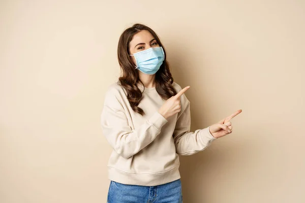 Glad leende kaukasiska kvinna bär medicinsk ansiktsmask från coronavirus pandemi, pekar finger höger, visar reklam, beige bakgrund — Stockfoto