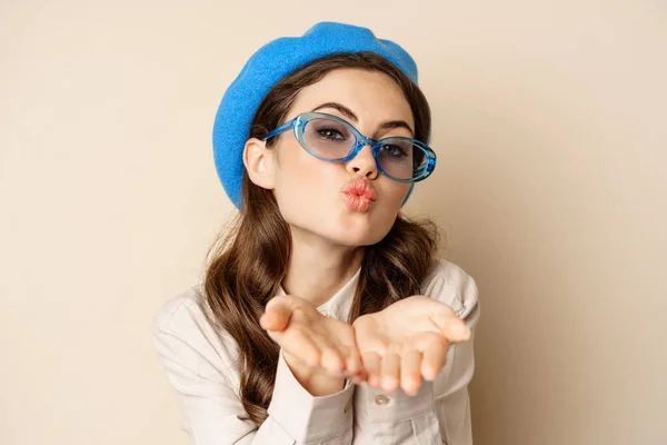 Beautiful stylish woman in sunglasses, blowing air kiss on hands near lips, gazing softly and flirty at camera, posing against beige background — Zdjęcie stockowe