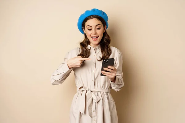 Портрет стильной современной женщины в верхней одежде, указывающей на экран мобильного телефона и выглядящей счастливой, улыбающейся довольной, стоящей на бежевом фоне — стоковое фото