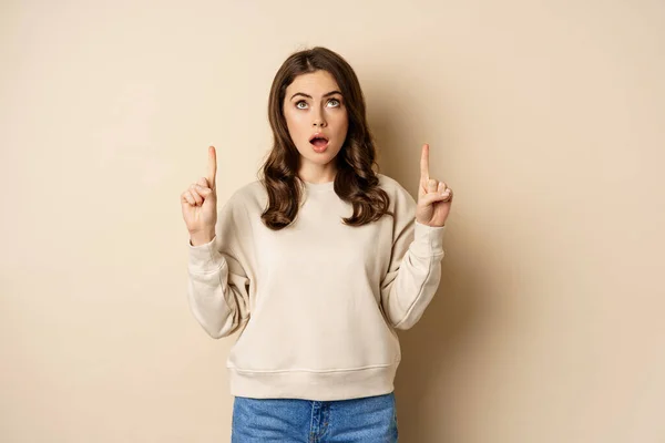 Ragazza stupita guardando banner annuncio, la lettura di testo promozionale verso l'alto, puntando il dito verso l'alto, in piedi su sfondo beige — Foto Stock