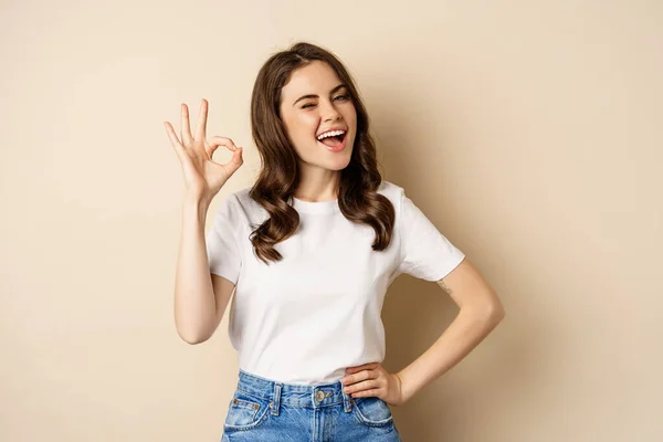 Stylish young woman smiling satisfied, showing okay, ok sign in approval, recommending, standing over beige background —  Fotos de Stock