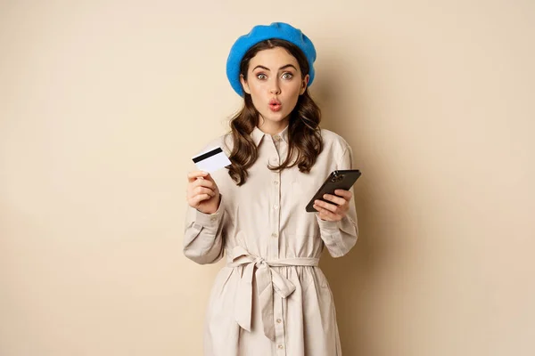 Портрет девушки, выглядящей взволнованной, скажем, вау, держа кредитку и мобильный телефон, стоя на бежевом фоне — стоковое фото