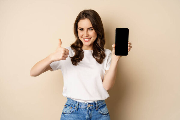 Image of happy young woman showing thumbs up and mobile phone screen, recommending webstore, online shopping application, standing pleased over beige background
