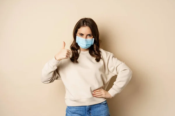 Covid-19, pandemic and quarantine concept. Young woman wears medical face mask during coronavirus omicron outbreak, showing thumbs up, standing over beige background — стоковое фото