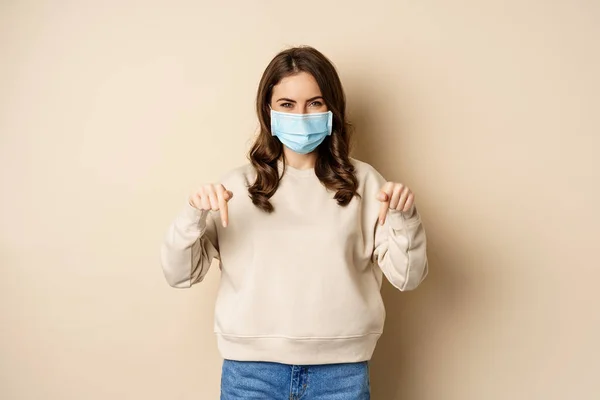 Mujer linda alegre en máscara facial médica, señalando con los dedos hacia abajo, mostrando publicidad, utilizando la protección de covid-19, fondo beige —  Fotos de Stock