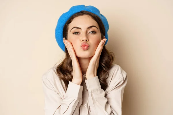 Image of stylish modern girl with makeup, clear glowing skin, pucker lips kissing face, standing over beige background coquettish — Zdjęcie stockowe