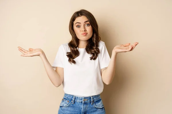 Young confused woman shrugging shoulders and with clueless face expression, standing over beige background — Stock Photo, Image