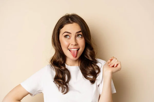 Close up of happy, carefree beautiful woman posing silly, showing tongue, having fun, standing in casual white t-shirt against beige background — стоковое фото