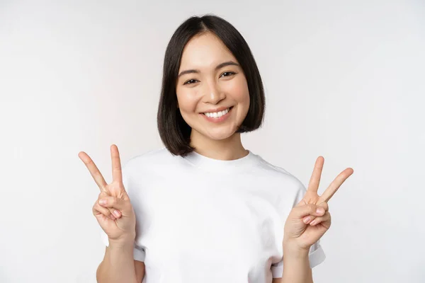 Bonito ásia menina mostrando paz, v-sign, sorrindo e olhando feliz para câmara, vestindo branco tshirt, estúdio fundo — Fotografia de Stock