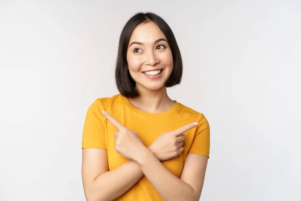 Söt asiatisk flicka pekar finger i sidled, visar vänster och höger promo, två val, varianter av produkter, stående i gul t-shirt över vit bakgrund — Stockfoto