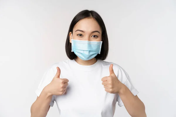 Covid-19, concepto de salud y personas. Joven mujer coreana feliz en máscara médica, mostrando los pulgares hacia arriba, de pie en camiseta sobre fondo blanco —  Fotos de Stock