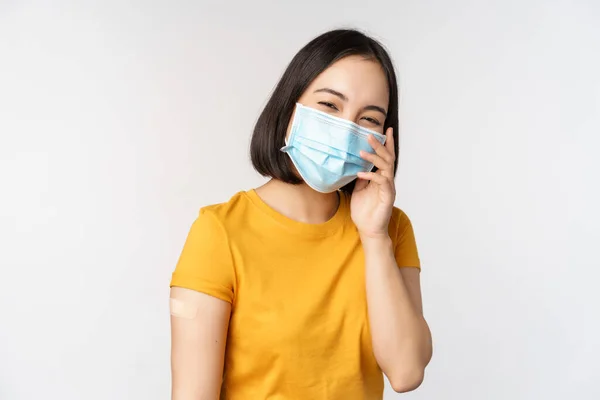 Covid-19, aşı ve sağlık konsepti. Tıbbi maskeli tatlı Asyalı kız, Coronavirus aşısından sonra yara bandı gösteriyor, beyaz arka planda duruyor. — Stok fotoğraf