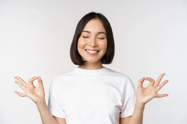 Porträtt av ung asiatisk kvinna mediterar, ler nöjd och utövar yoga, blunda och meditera, andas lugn, står över vit bakgrund — Stockfoto