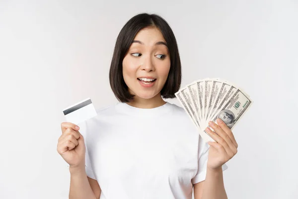 Porträt einer asiatischen Frau mit Geld, Dollars und Kreditkarte, die beeindruckt und erstaunt aussieht und im T-Shirt vor weißem Hintergrund steht — Stockfoto