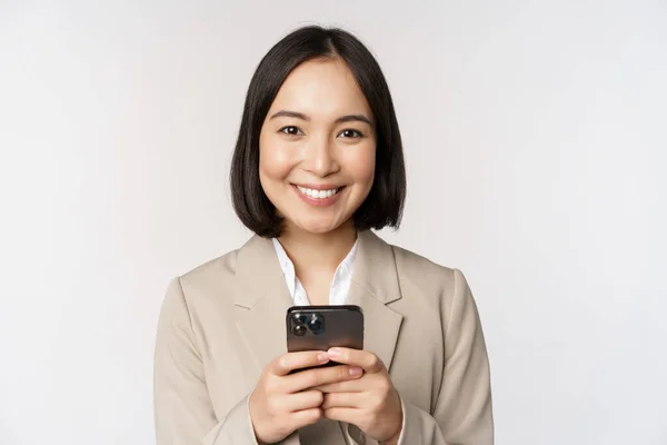 Image d'une femme d'affaires asiatique en costume, tenant un téléphone mobile, utilisant une application pour smartphone, souriant à la caméra, fond blanc — Photo