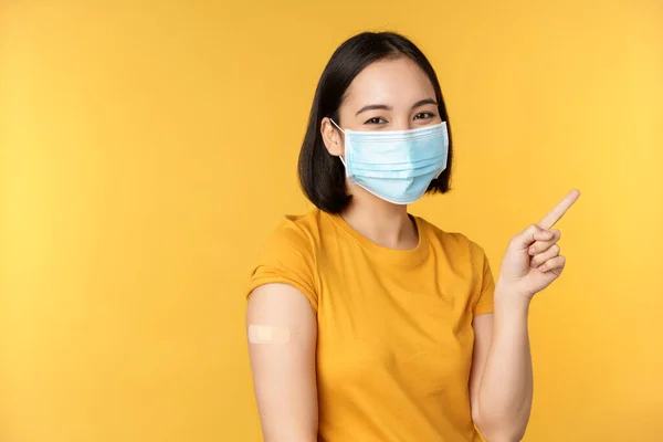 Vacinação a partir de covid e conceito de saúde. Imagem da menina coreana sorridente em máscara facial médica, ajuda banda no ombro, apontando o dedo para a propaganda banner, fundo amarelo — Fotografia de Stock
