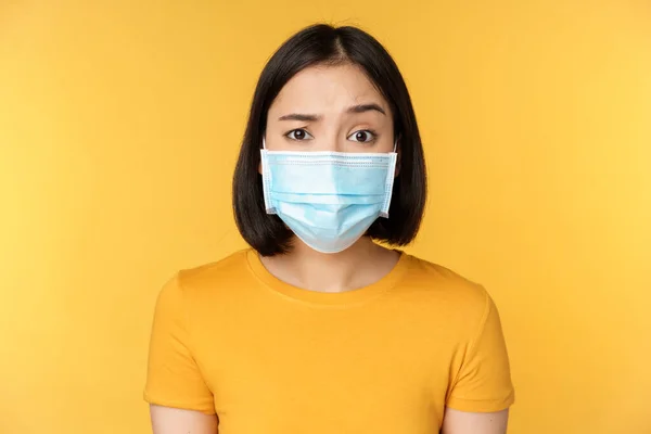 Portrait of skeptical and confused asian woman in medical face mask, raising eyebrow doubtful, standing over yellow background — Stock Photo, Image