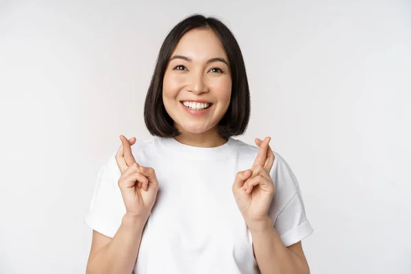 Esperançoso asiático menina cruz dedos, fazendo desejo, antecipando, desejando smth, de pé em tshirt sobre branco fundo — Fotografia de Stock