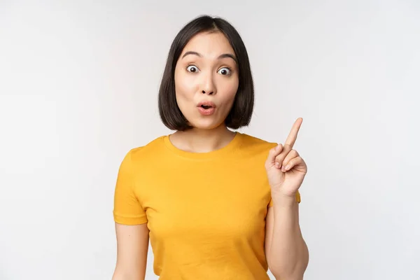 Gyönyörű fiatal ázsiai nő mutogatott ujjal felfelé, mosolygott és nézett szórakoztató kamera, bemutató reklám, bejelentés a tetején, fehér háttér — Stock Fotó