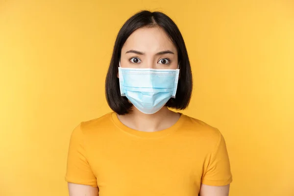 Portrait of shocked asian woman looking concerned and startled at camera, wearing covid-19 medical face mask, standing against yellow background — Stock Photo, Image