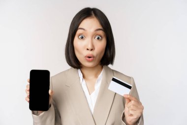 Mutlu yüzlü, mutlu bir kadın, kredi kartı ve akıllı telefon uygulaması ekranı gösteriyor, beyaz arka planda takım elbise içinde duruyor.