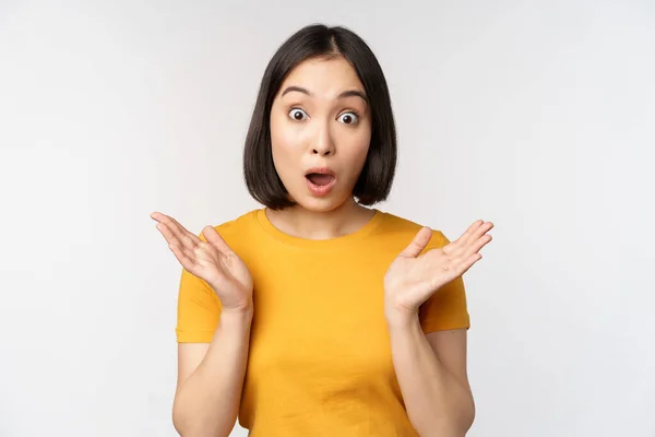 Primer plano retrato de mujer asiática mirando sorprendido, wow cara, mirando impresionado a la cámara, de pie sobre fondo blanco en camiseta amarilla — Foto de Stock