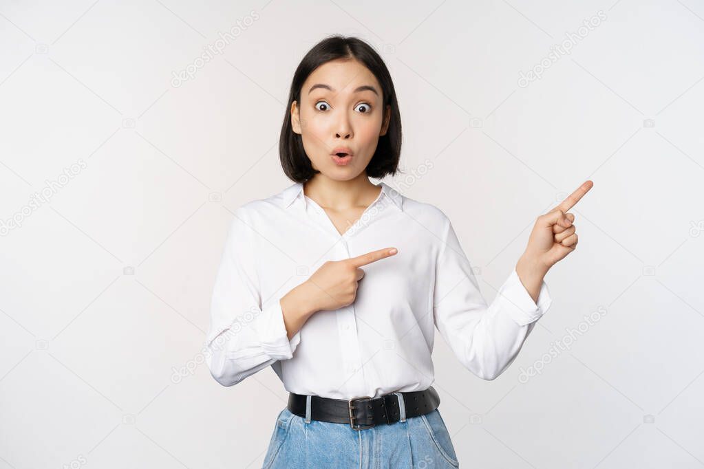 Surprised young woman pointing fingers right. Asian girl showing banner and looking enthusiastic, interested in advertising, standing over white background