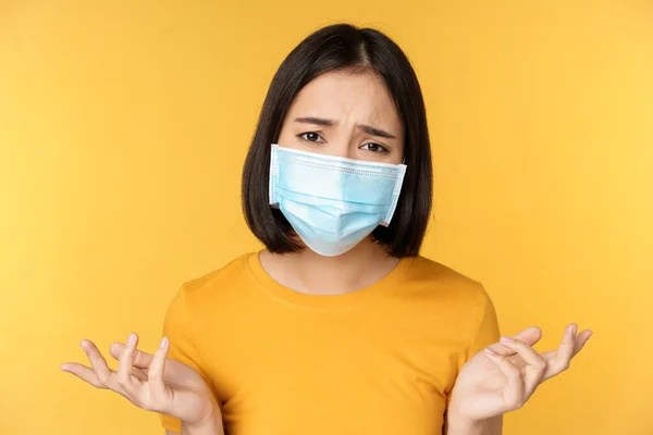 Close up retrato de mulher asiática confusa em máscara facial médica, ombros encolhendo os ombros e olhando perplexo, de pé contra fundo amarelo — Fotografia de Stock