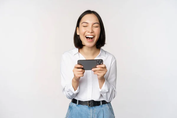 Изображение молодой азиатки, смотрящей на приложение смартфона, держащей мобильный телефон и смотрящей на экран, смеющейся и улыбающейся, стоящей на белом фоне — стоковое фото