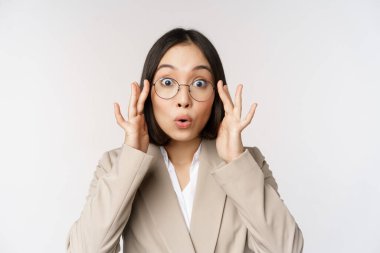 Gözlüklü Asyalı iş kadını portresi, kameraya şaşırmış bakıyor, beyaz arka planda takım elbise içinde duruyor.