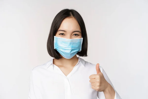 Concepto de covidio y salud. Retrato de mujer asiática usando mascarilla médica y mostrando los pulgares hacia arriba, de pie sobre fondo blanco —  Fotos de Stock