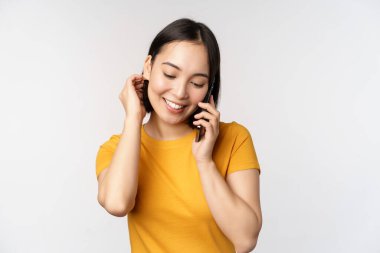 Romantik ve sevimli Asyalı kız telefona cevap veriyor, cep telefonuyla konuşuyor, gülümsüyor, sarı tişörtle beyaz arka planda dikiliyor.