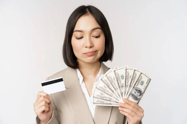 Pengusaha wanita yang bijaksana, korea perusahaan wanita menunjukkan kartu kredit dan uang tunai, dolar di tangan, berdiri di atas latar belakang putih dan berpikir — Stok Foto