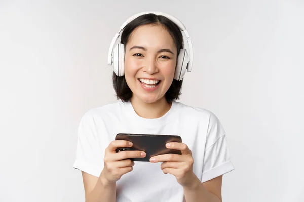 Joyeux asiatique fille gamer, jouer sur téléphone mobile, regarder sur smartphone, porter des écouteurs, debout sur fond blanc — Photo