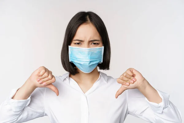 Retrato de ásia mulher no médico rosto máscara mostrando polegares para baixo com decepcionado, cansado rosto expressão de pé sobre branco fundo — Fotografia de Stock