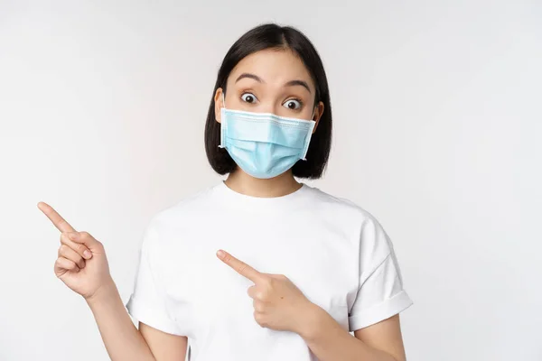 驚いたアジアの少女は 医療マスクで 指差し指を左に プロモーションを提供し 眉を上げ 驚きの反応を示し 白い背景に立って — ストック写真
