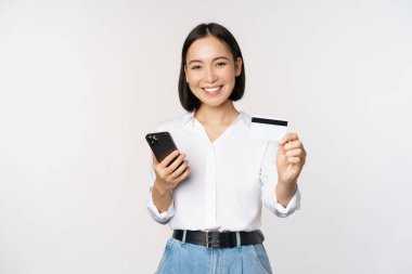 İnternetten alışveriş konsepti. Gülümseyen modern Asyalı kız kredi kartını gösteriyor, cep telefonunu tutuyor, akıllı telefonuyla sipariş veriyor, beyaz arka planda duruyor..